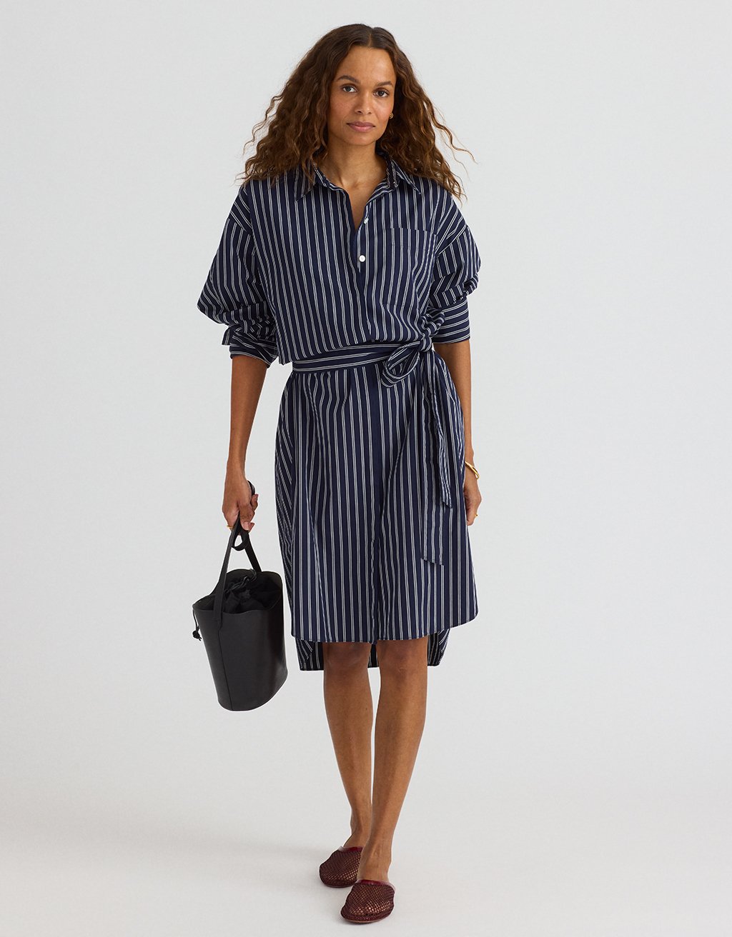 Woman wearing a navy and white striped shirtdress holding a black handbag on a plain background