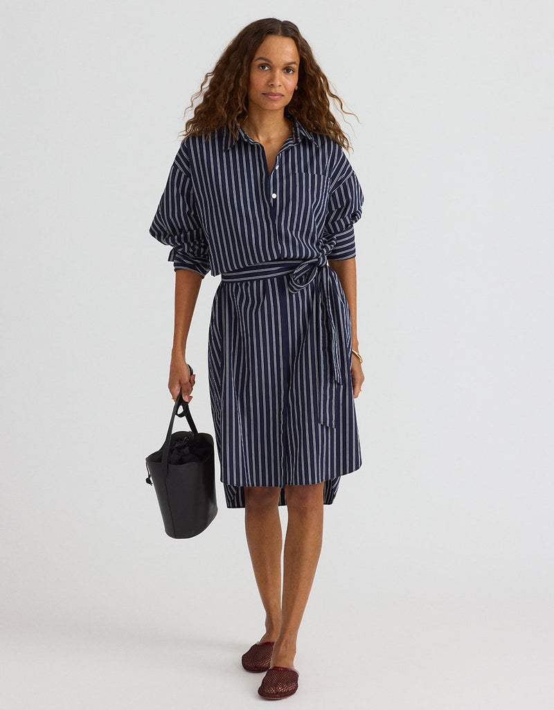 Woman wearing a navy and white striped shirtdress holding a black handbag on a plain background