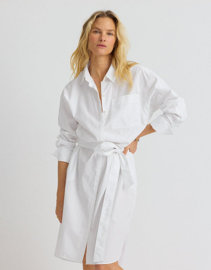 Woman wearing a belted white shirtdress on a plain background