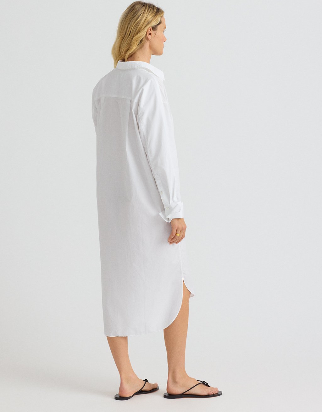 Woman wearing a white shirtdress and black sandals on a plain background