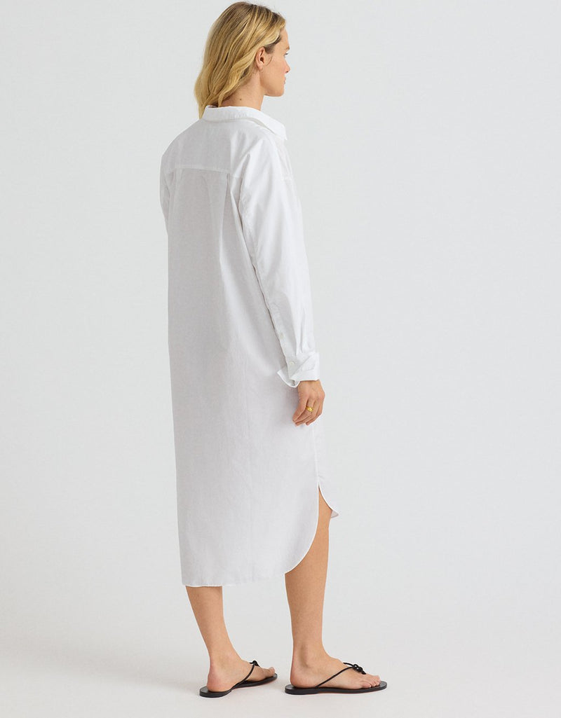 Woman wearing a white shirtdress and black sandals on a plain background