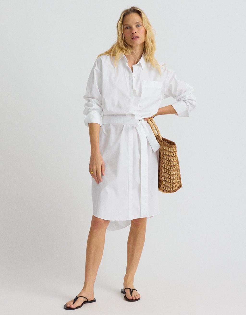 Woman wearing a belted white shirtdress and black sandals holding a woven bag on a plain background