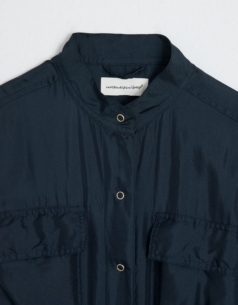 Close-up of navy silk utility jumpsuit with a brand label and gold snap buttons on a light gray background