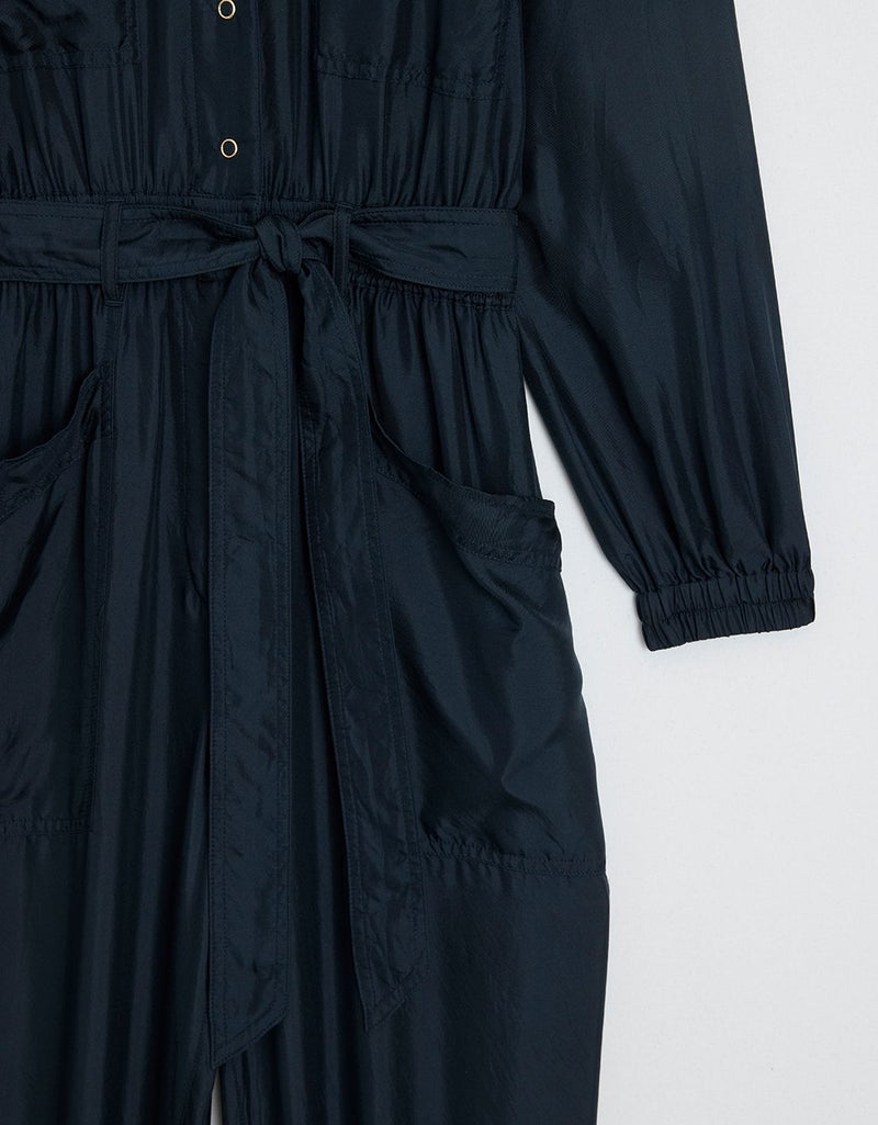Detail close-up featuring ruched accents, elasticized sleeve cuffs, tie waist belt and side pockets of a navy silk utility jumpsuit on a light gray background