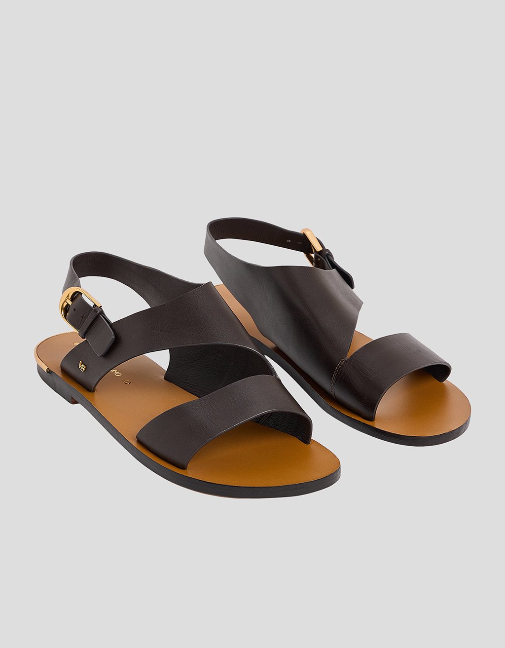 Side view of a brown leather sandal with a gold buckle ankle strap on a light gray background